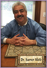 The image shows a man seated at a desk with a nameplate that reads Dr. Samer Alois. He is smiling, wearing glasses, and has his hands crossed on his lap. Behind him, there s a framed certificate on the wall above the desk.