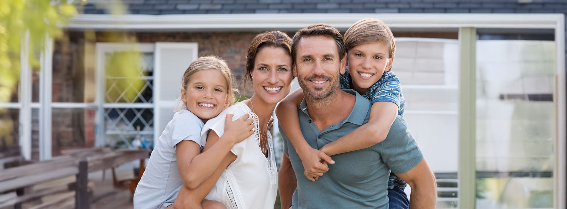 This is a photograph of a family of four, including two adults and two children, posing for a picture outdoors with a residential house in the background.
