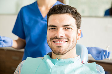 The image shows a man sitting in a dental chair with a smile, wearing a blue surgical mask and a white apron, accompanied by a female dental professional standing behind him.